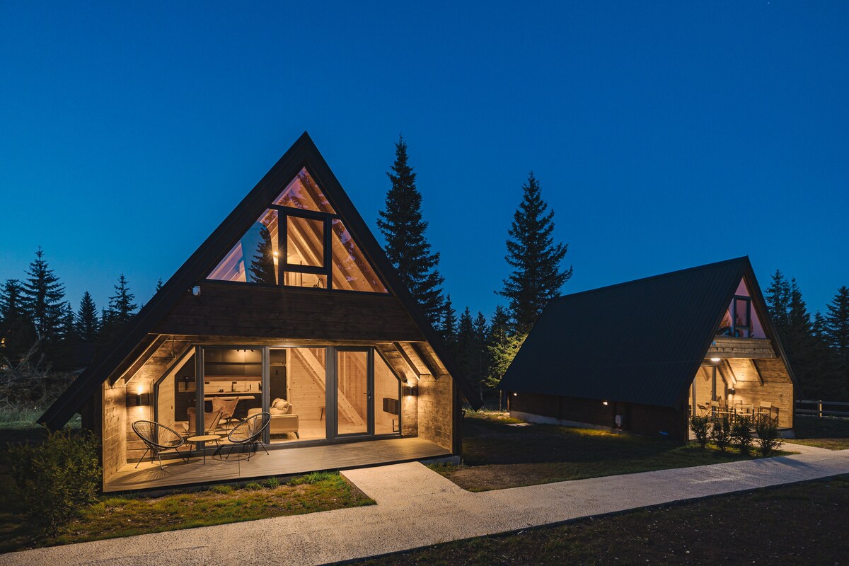 Two chic villas are shown under a dusk sky, featuring A-frame designs with wooden exteriors and large glass doors. Warm lighting enhances their architectural details, and surrounding pine trees add to the serene ambiance, with a well-maintained pathway leading to the entrances.