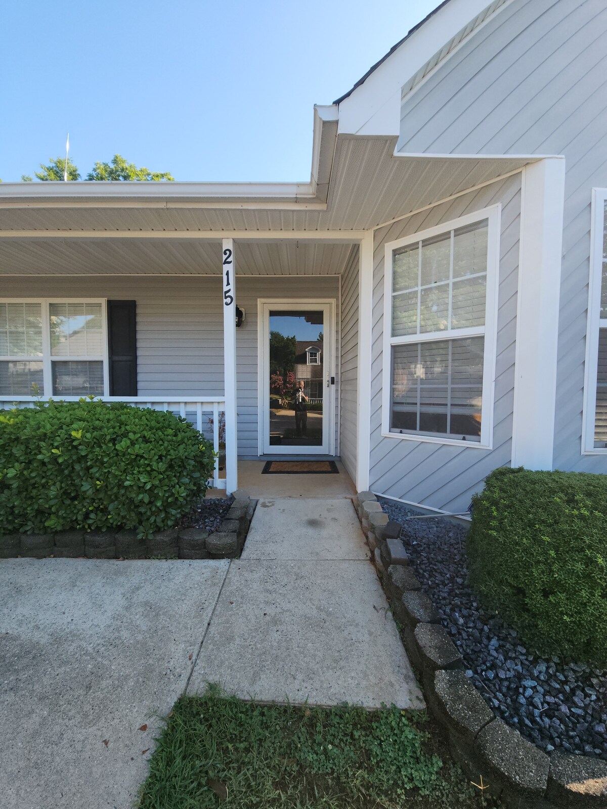 The entrance features a small pathway leading to a front door framed by large windows. Lush green shrubs line the pathway, and a covered porch provides a welcoming space that is well-lit by natural sunlight.