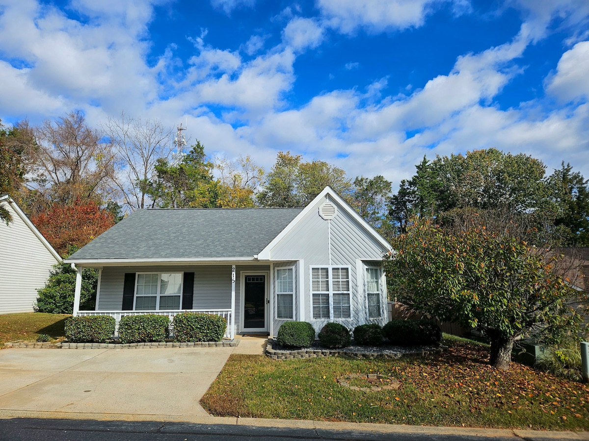 A cozy home presents a welcoming façade with a light grey roof and white siding. Vibrant greenery frames the entrance, along with neatly trimmed bushes. The front porch features black shutters and a charming entryway, set against a backdrop of a clear blue sky with wispy clouds.