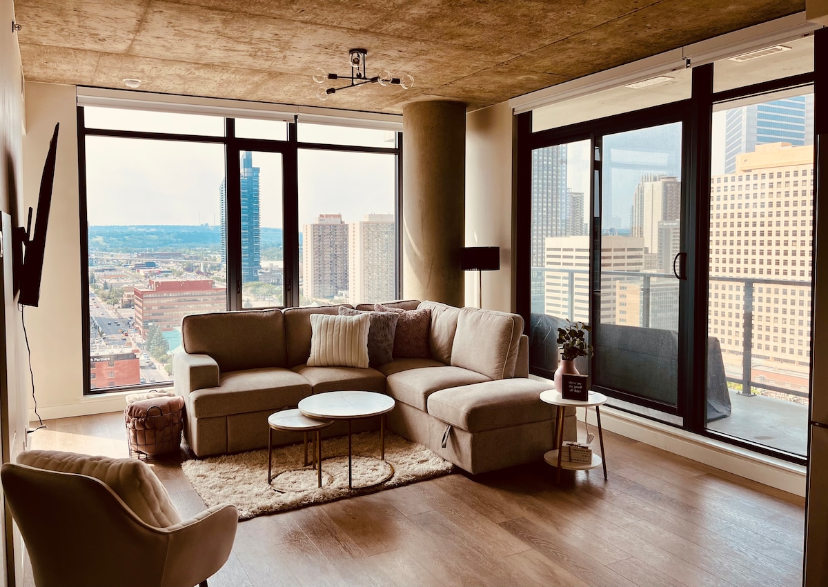 A modern living area features a spacious sectional sofa, elegantly arranged around a round coffee table. Floor-to-ceiling windows present panoramic views of the urban landscape, complemented by natural light streaming into the room. A cozy area rug adds warmth to the hardwood flooring.