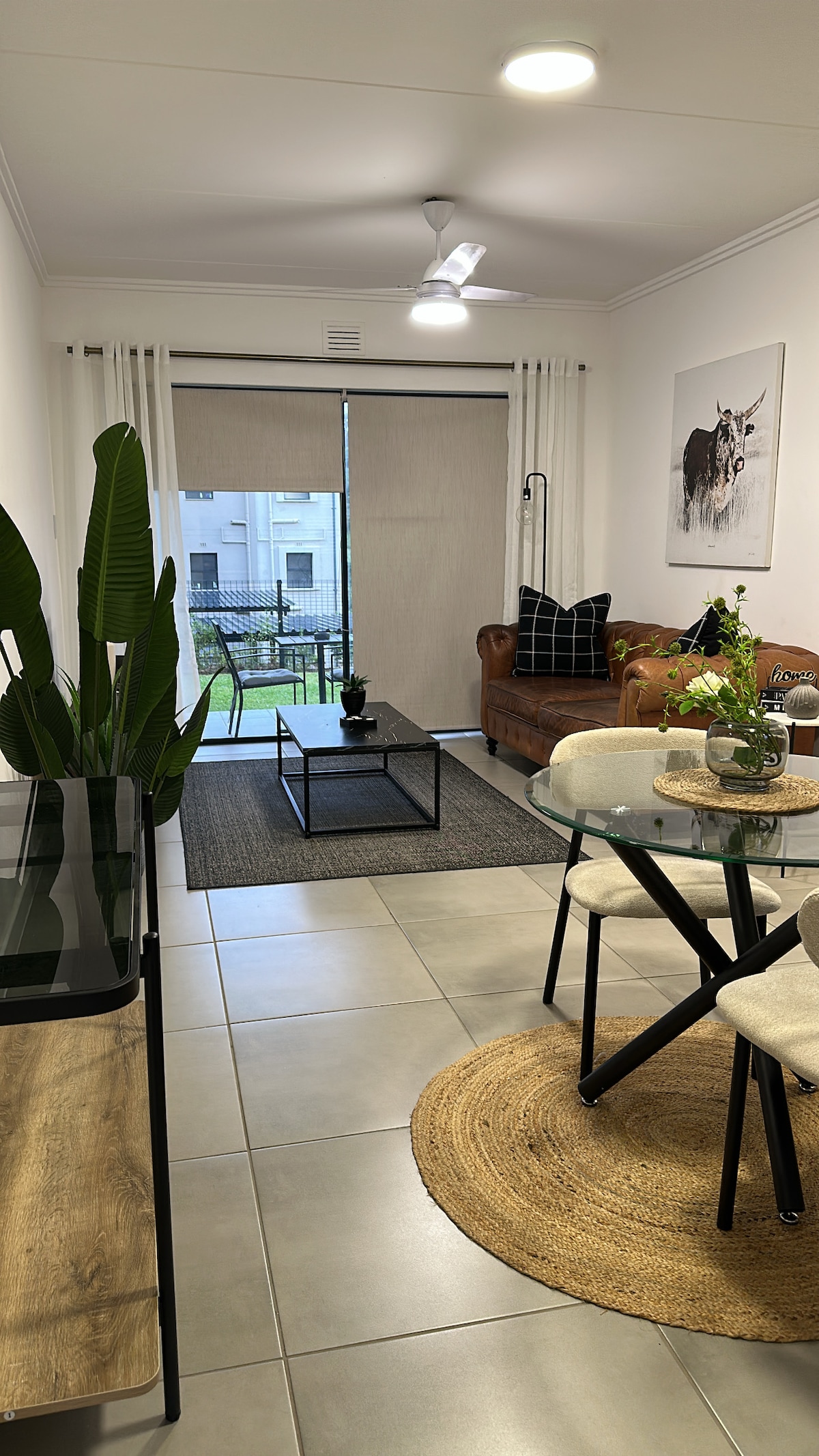 A modern living area features a brown leather couch paired with a patterned cushion, alongside a sleek glass coffee table set on a textured rug. A dining table with light-colored chairs sits nearby, while large windows offer a view of the outdoor space, allowing natural light to fill the room.