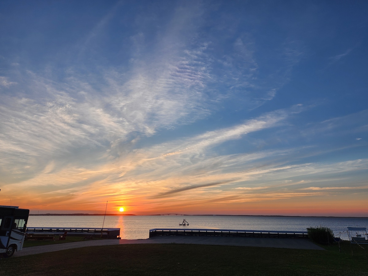 A serene view of a sunset over the water is captured, where the sky is filled with soft hues of orange, pink, and blue. Calm waves can be seen reflecting the diminishing light as silhouettes of distant boats rest on the horizon.