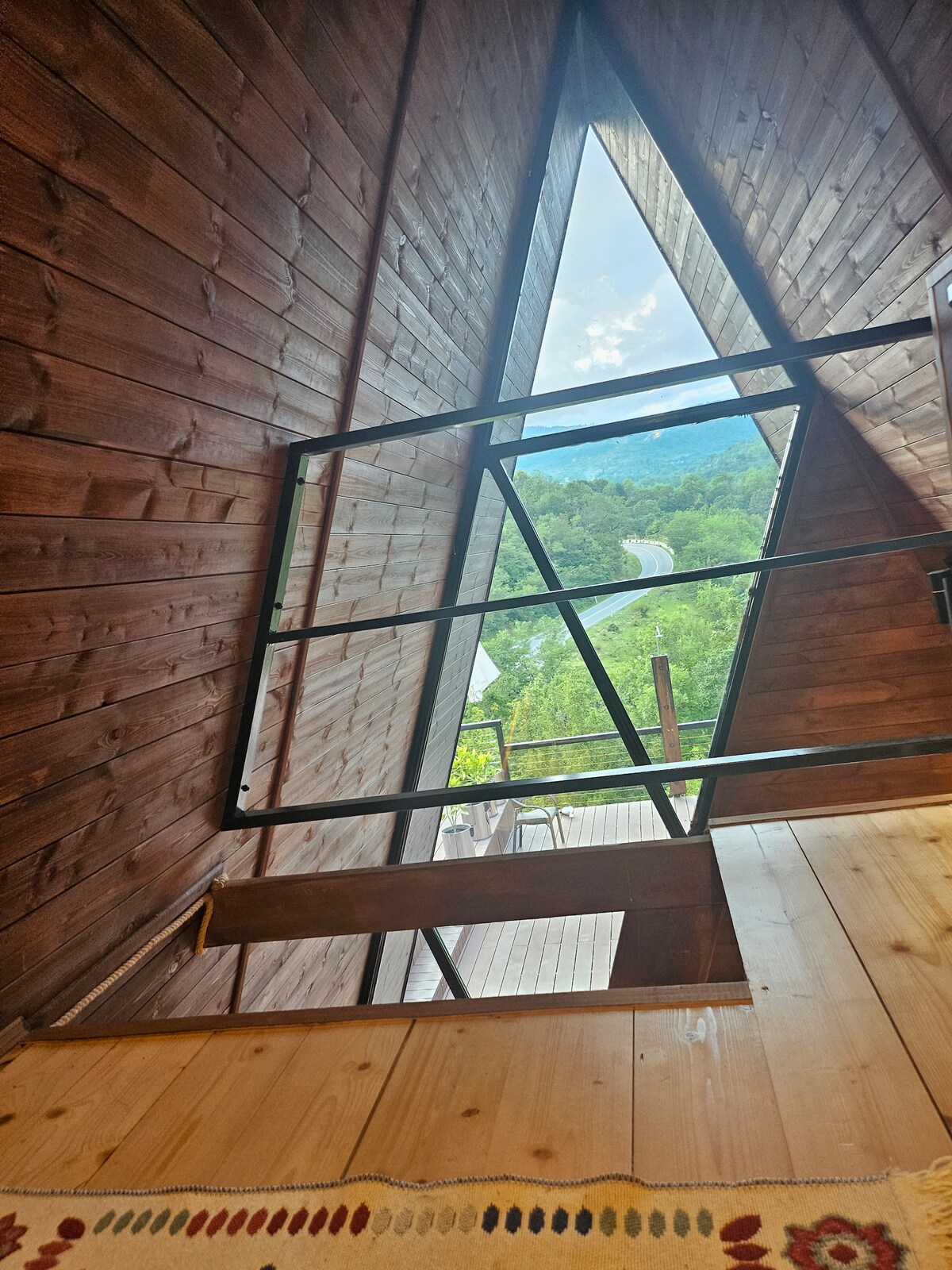 A wooden interior with sloped A-frame walls features large windows that offer a view of the surrounding greenery and winding path. Natural light illuminates the staircase, which leads to a spacious outdoor terrace, enhancing the connection with nature.