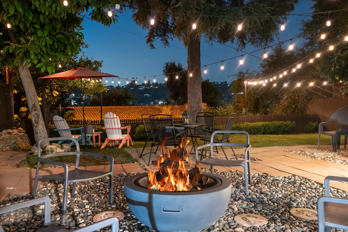 An outdoor space is illuminated by string lights overhead, creating a warm ambience. A fire pit sits at the center, surrounded by several seating options, including metal chairs and colorful lounge chairs. Lush greenery and distant city views add to the serene atmosphere.
