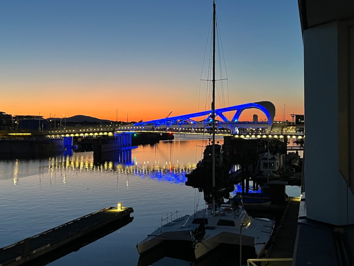 The image captures a serene waterfront scene at dusk, featuring a modern bridge illuminated in blue hues. The calm water reflects the vibrant orange and blue sky, creating a tranquil atmosphere. Sailboats are visible moored along the dock, enhancing the picturesque view.
