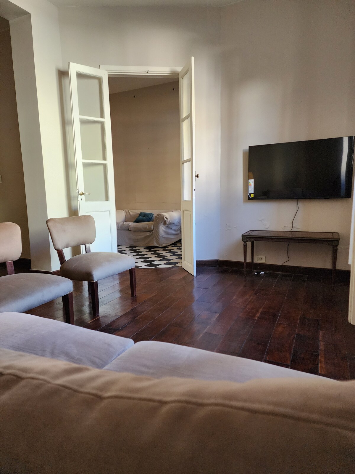A light-filled living area features two armchairs and a sofa, overlooking an adjacent room with a checkerboard floor. Double doors stand open, connecting the spaces. A flat-screen television is mounted on the wall, providing an inviting setup for relaxation.