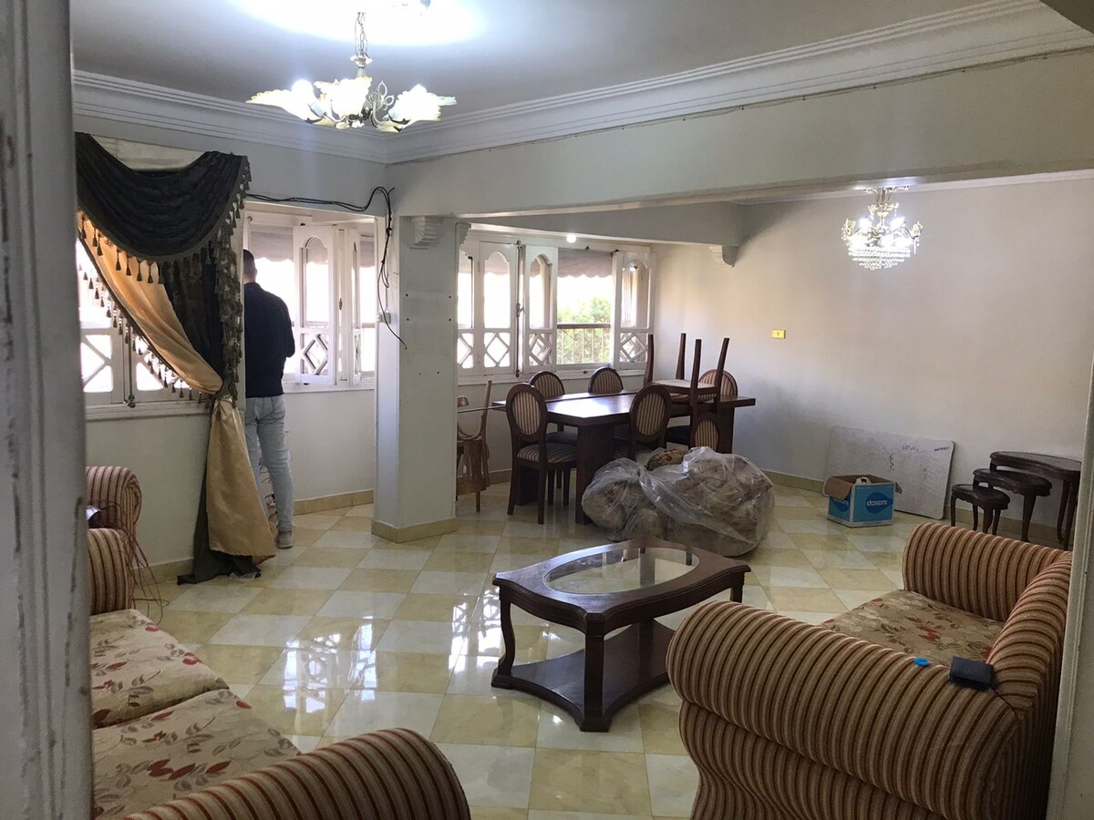 The spacious common area features a dining table with six chairs, complemented by a glass coffee table set on a tiled floor. Two striped sofas are arranged nearby, and large windows provide ample natural light, framed by elegant drapes.