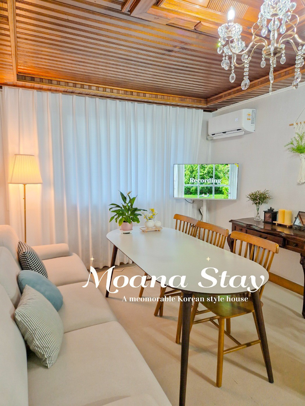 A cozy living area features a light-colored sofa adorned with decorative cushions. A wooden dining table is set for six, complemented by a stylish cabinet and a potted plant. Soft ambient lighting is provided by a floor lamp, with large curtains framing a window.