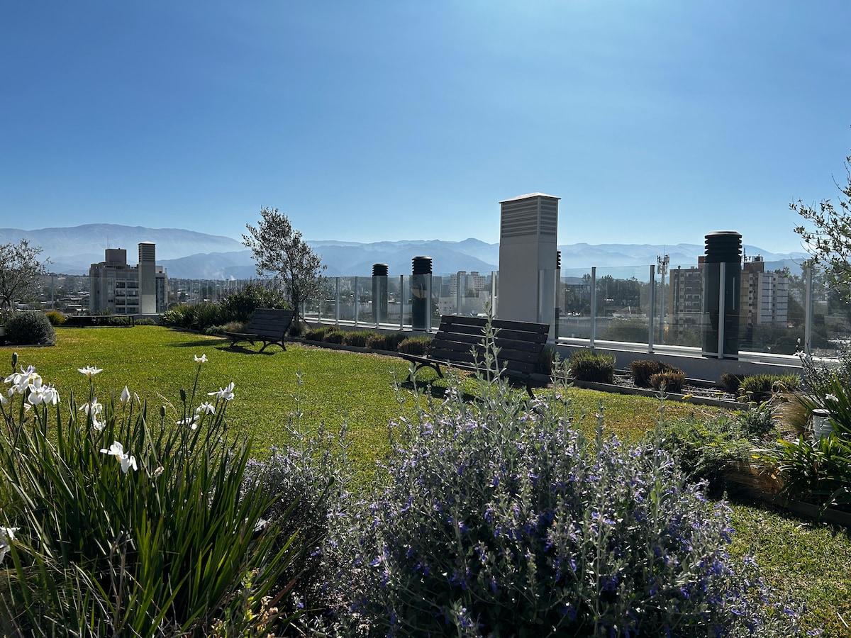 A green rooftop terrace is visible, adorned with lush grass and flowering plants. Seating areas are provided along the edge, with expansive views of the surrounding city and mountains under a clear blue sky.