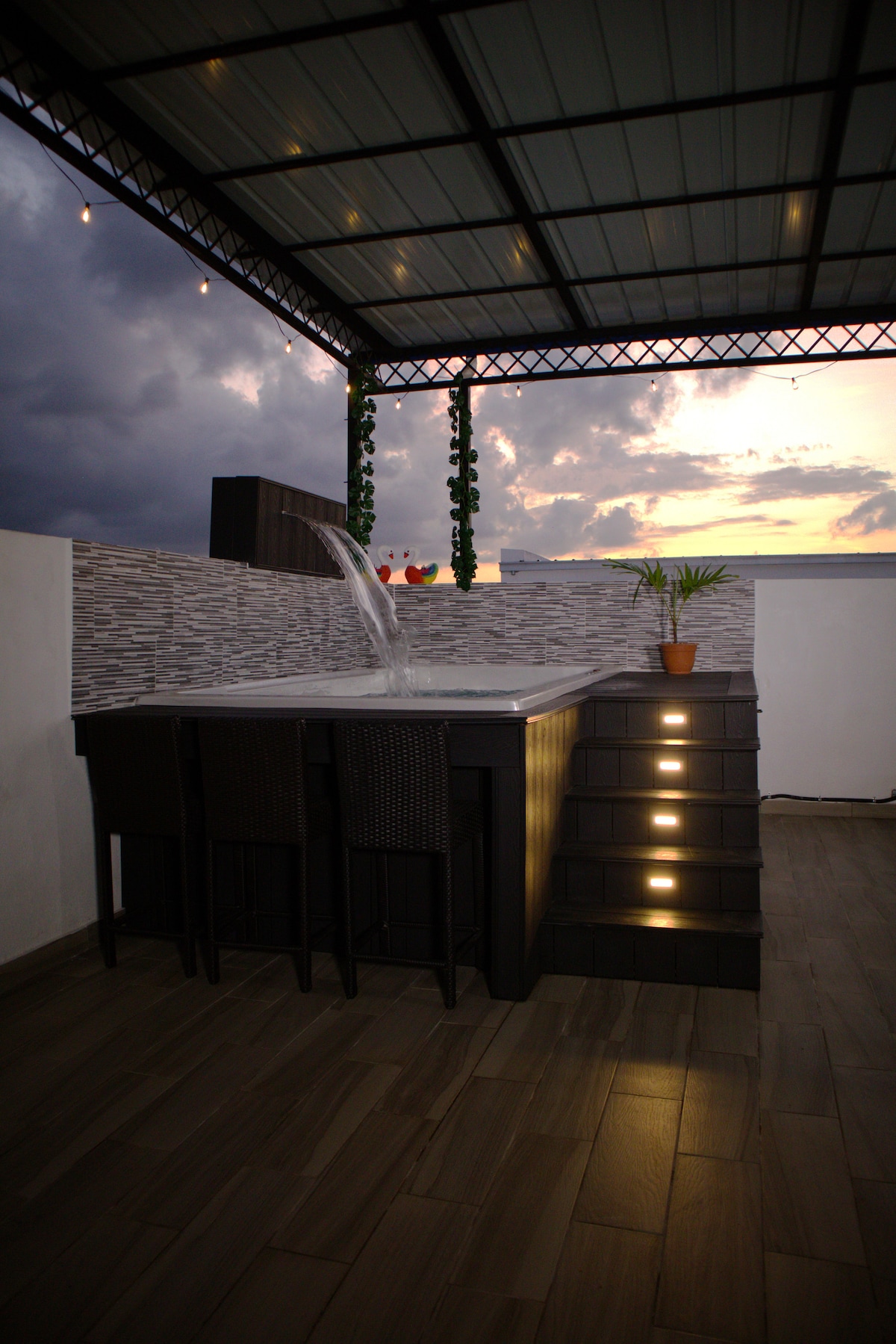 An outdoor seating area features a stone bar coupled with high-backed chairs, creating a space for relaxation. Water gently flows from a stylish fountain, while soft lighting and greenery add an inviting touch. A vibrant sunset sky provides a picturesque backdrop.