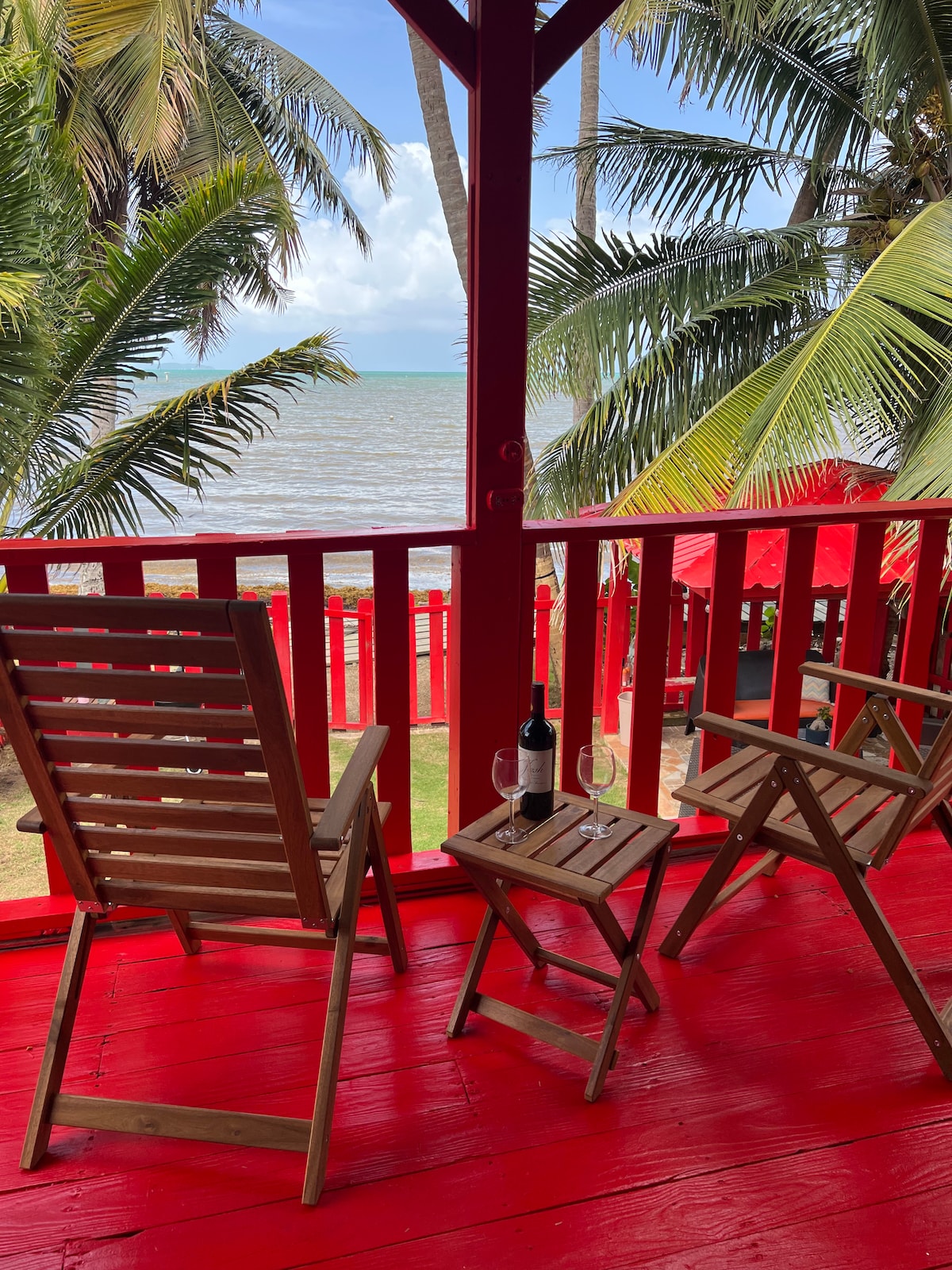 A shaded outdoor balcony features two wooden folding chairs positioned around a small table. A bottle of wine and two glasses rest on the table, with tropical palm trees and the ocean visible in the background.
