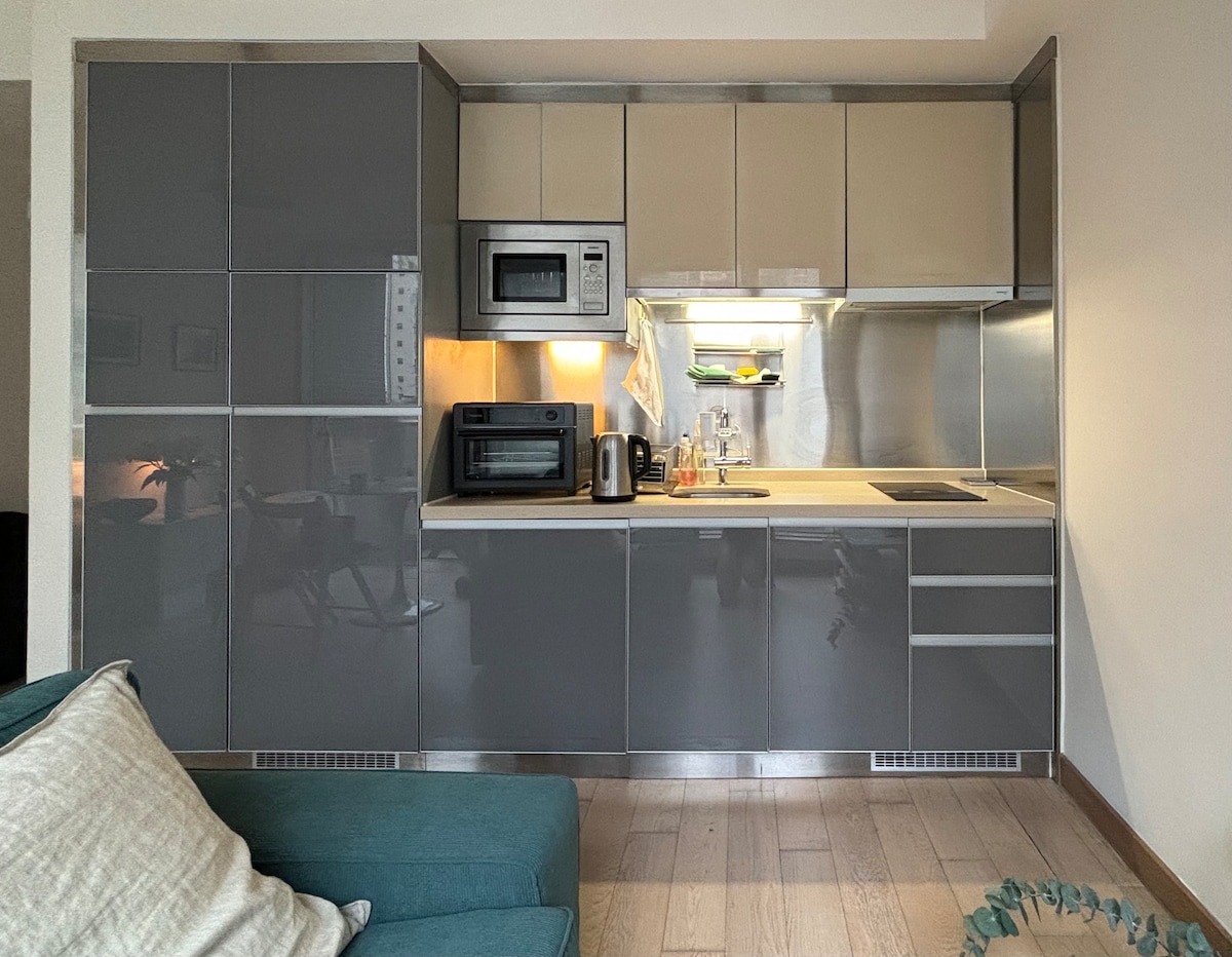 The kitchen area features sleek gray cabinetry and modern appliances, including a microwave, coffee maker, and electric kettle. A stainless steel countertop is neatly arranged with utensils, providing a functional cooking space that is well-lit for convenience.