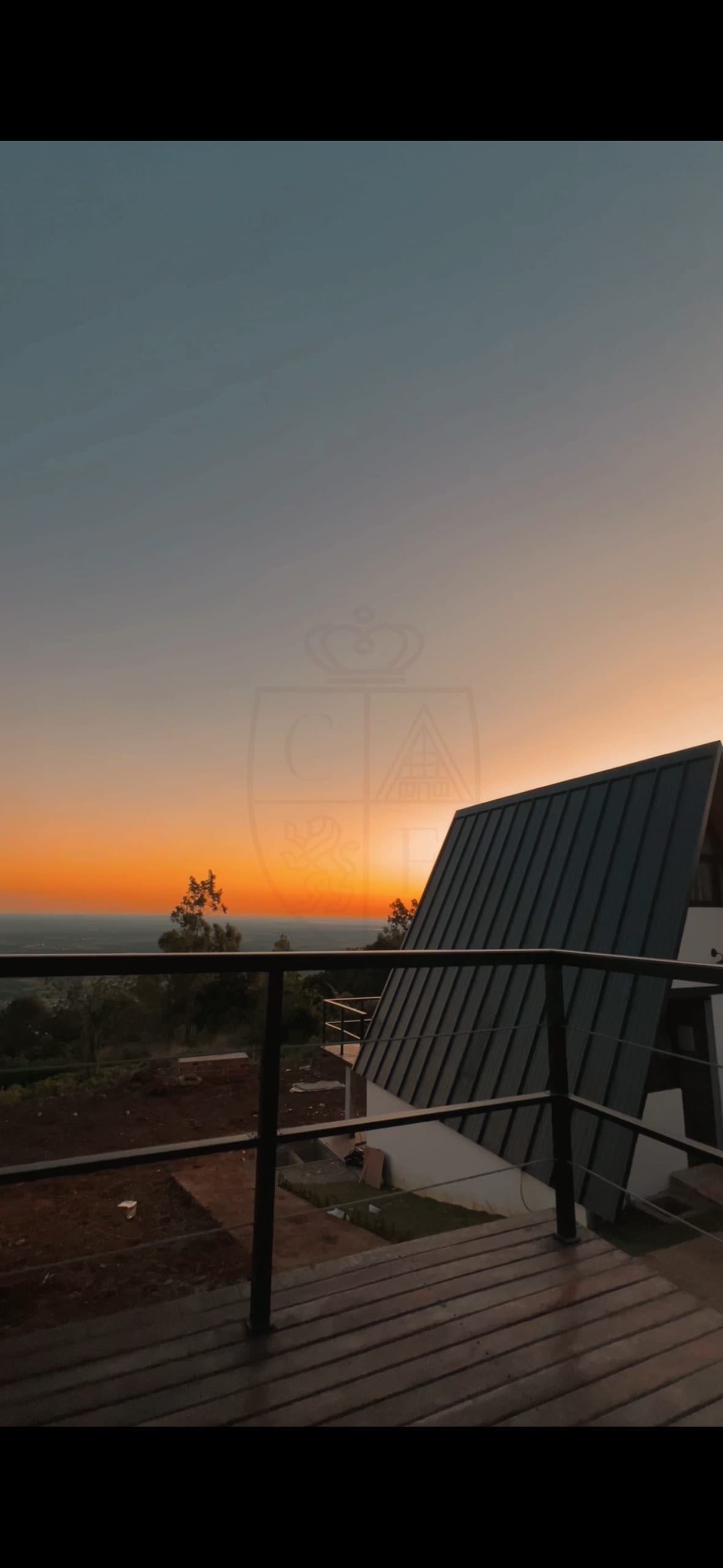 A scenic sunset is captured with vibrant hues of orange and blue over the horizon, viewed from a balcony. In the foreground, a modern structure is visible, with large windows reflecting the warm light, while greenery is seen in the background.