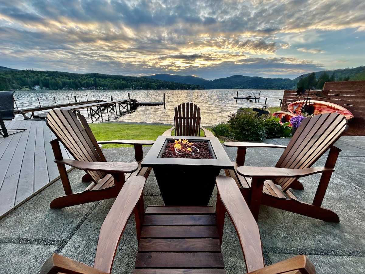 A serene outdoor seating area features four wooden Adirondack chairs arranged around a central fire pit. The setting showcases a calm lake with a picturesque sunset backdrop, while a dock and kayaks are visible in the distance, inviting guests to enjoy the tranquil atmosphere.