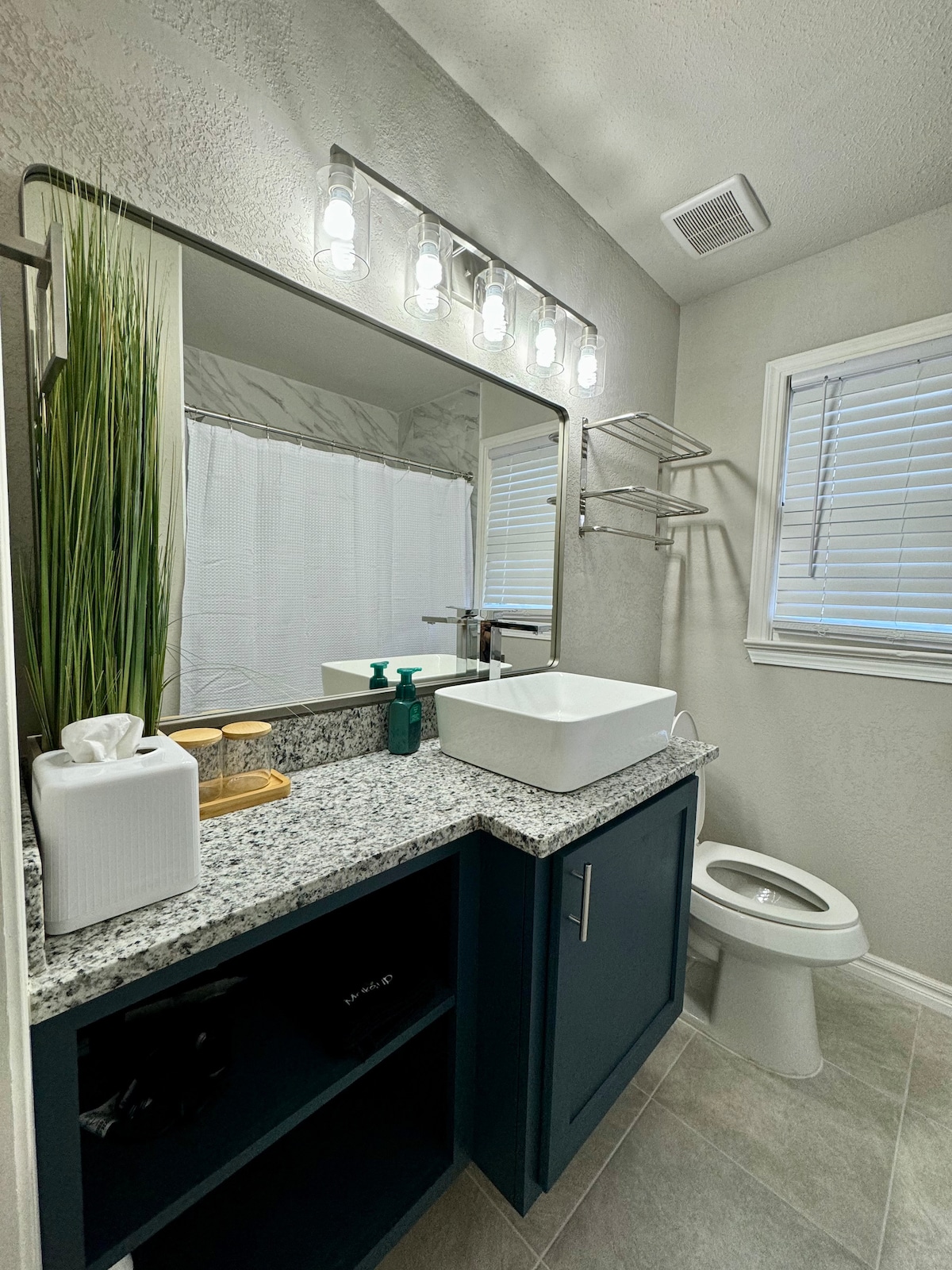 A modern bathroom features a stylish granite countertop with a square sink and a wall-mounted mirror above. Shelving holds bathroom essentials, and natural light filters through a window with blinds. The flooring is light-colored tile, and a white shower curtain is visible in the background.