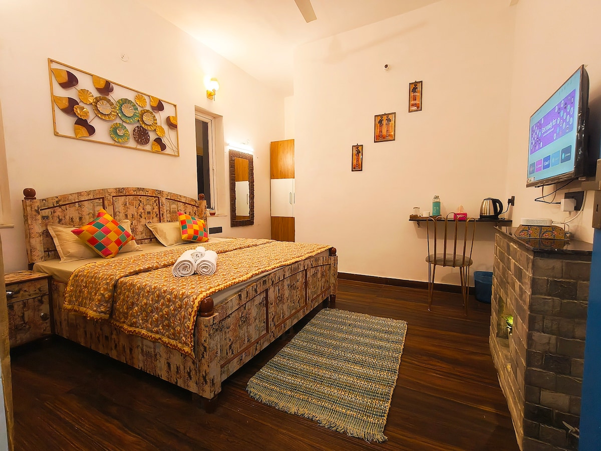 A spacious bedroom is showcased, featuring a well-made bed adorned with patterned linens and colorful cushions. A flat-screen TV is mounted on the wall, and a small table is positioned beside a wall mirror. Warm lighting enhances the cozy atmosphere.