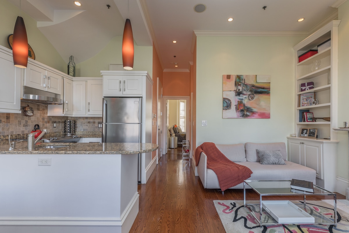 A kitchen area features granite countertops and stainless steel appliances. A modern sofa is positioned in a well-lit living space, with a colorful artwork displayed on the wall. The flooring is hardwood, enhancing the openness of the layout.