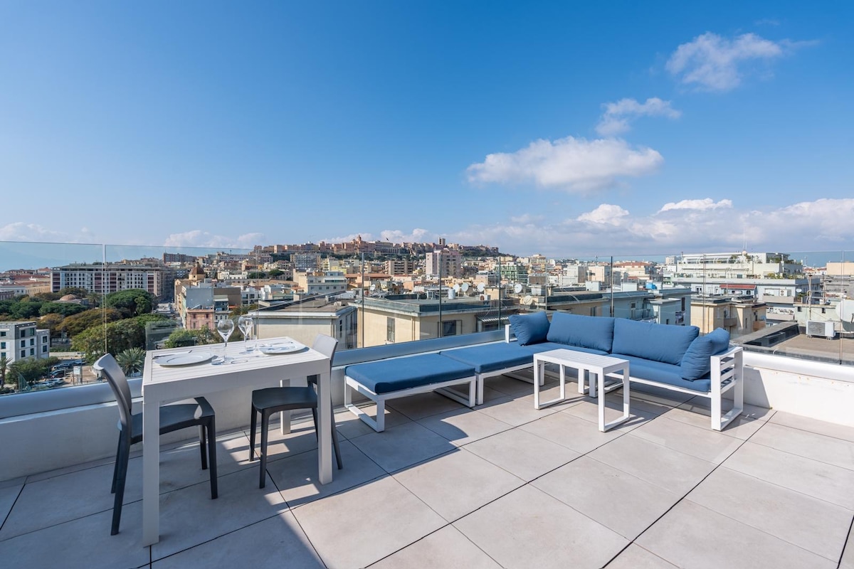 A private terrace is visible, featuring a dining table set for two and a lounge area with blue cushions. The space offers panoramic views of the city skyline, including historic buildings and surrounding landscapes under a clear blue sky.