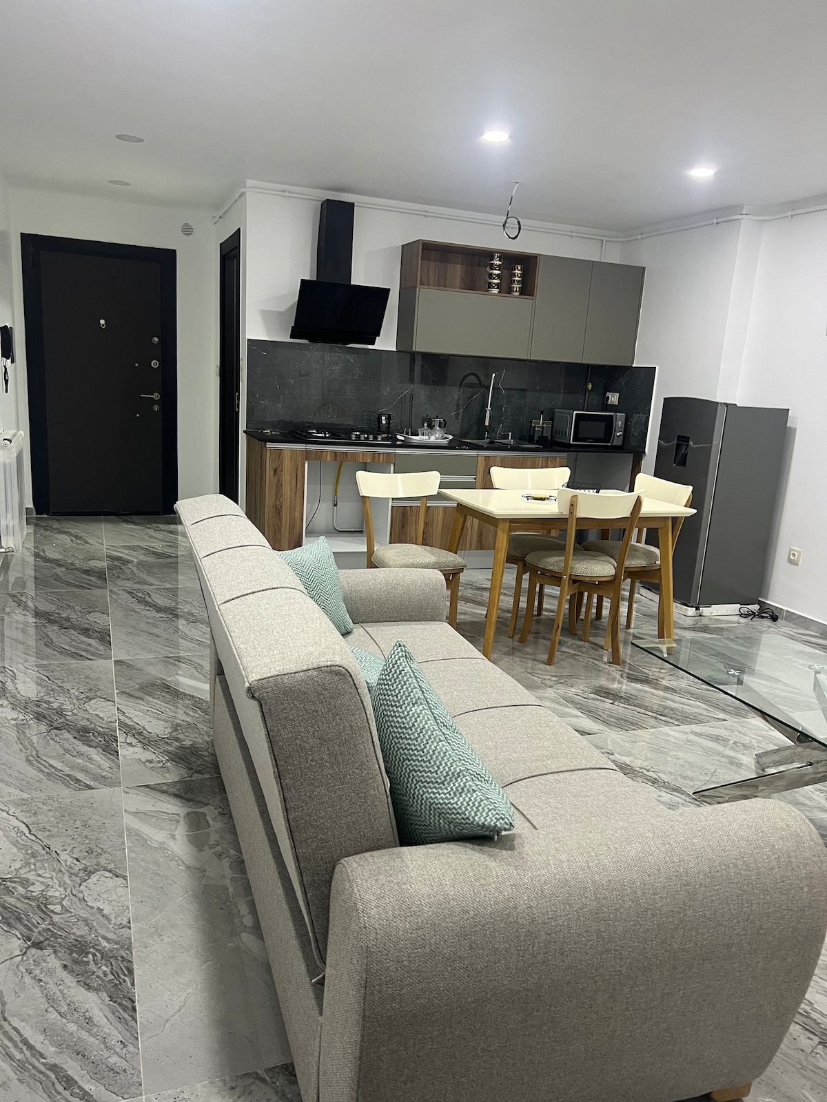 A modern kitchen and dining area are displayed, featuring a light wood dining table surrounded by four chairs. A comfortable sofa with accent cushions is positioned nearby. Sleek marble flooring adds to the contemporary aesthetic, complemented by a wall-mounted TV and stainless steel refrigerator.