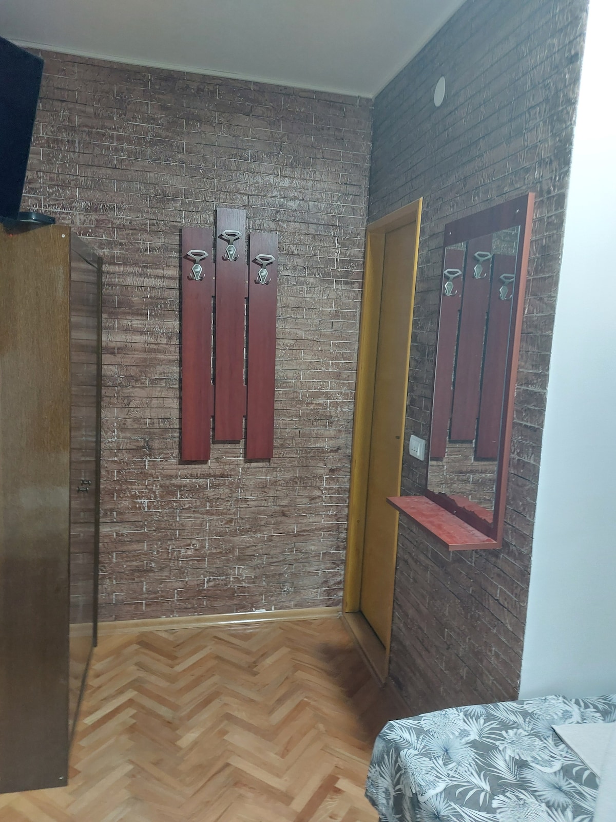 A narrow hallway features a textured brick-patterned wall and wooden flooring. A wooden wardrobe stands on the left, while a red wall-mounted coat rack is positioned above a small shelf. A mirror is attached to the wall, reflecting the hallway's design.