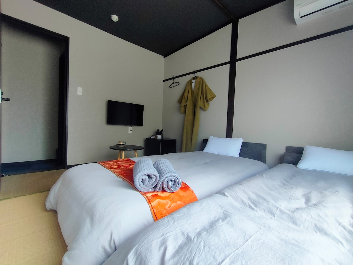 A cozy sleeping area features two spacious beds dressed in soft linens, with neatly rolled towels placed on a decorative fabric. A television is mounted on the wall, while a garment hangs nearby. The room is enhanced by neutral wall tones and organized furniture.