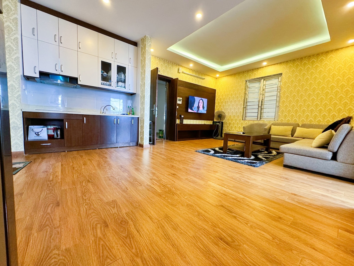 A spacious living area showcases a combination of modern and comfortable furnishings. A large, beige sofa is placed alongside a dark wooden coffee table. The kitchen area features white cabinets and appliances, while light-colored flooring enhances the room's openness. Natural light is visible through a window.