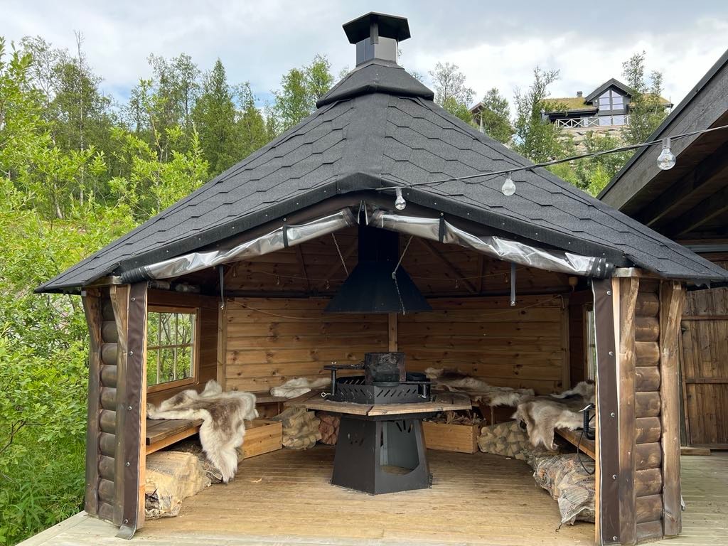 A spacious grill hut is featured, enclosed by wooden benches and decorated with warm fur throws. A central grill is positioned beneath a black chimney, providing a cozy gathering space. Surrounding greenery offers a natural backdrop for outdoor meals and socializing.