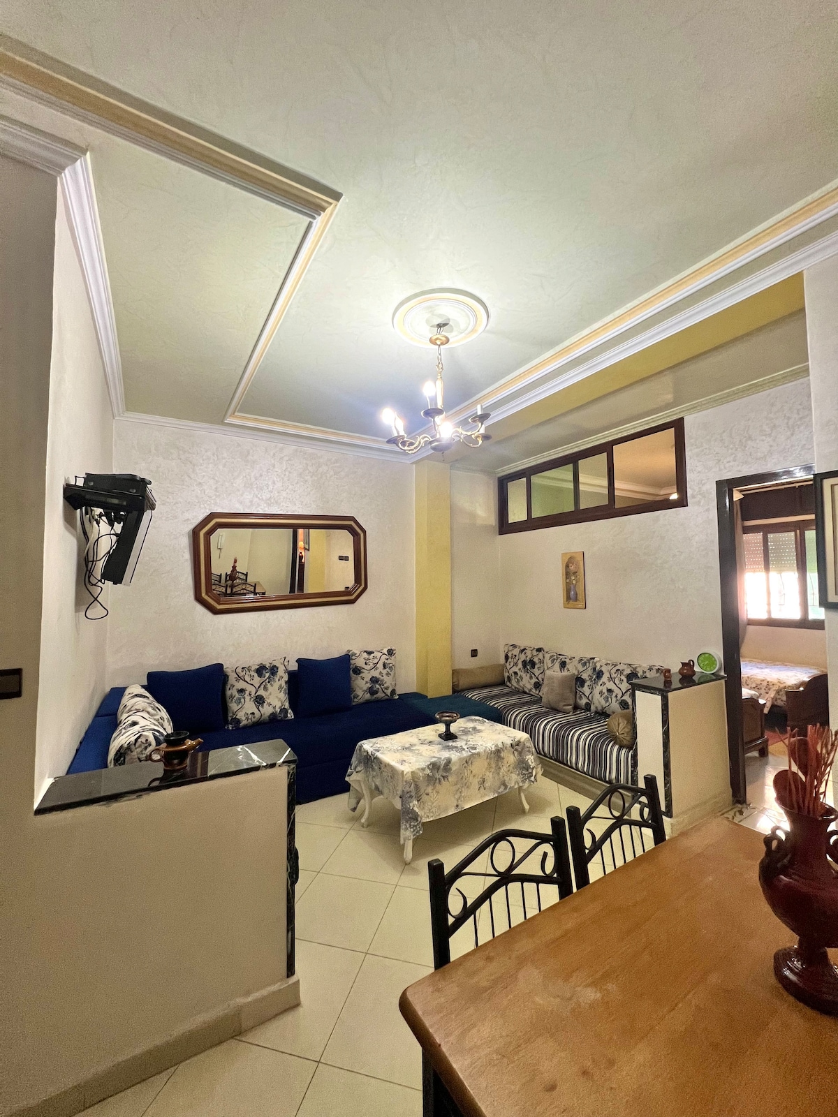 A welcoming living area features a blue sofa adorned with decorative pillows and a striped chair. A wooden dining table sits adjacent, with a mirror reflecting the room's light fixtures. Soft lighting enhances the pale walls and detailing of the ceiling.