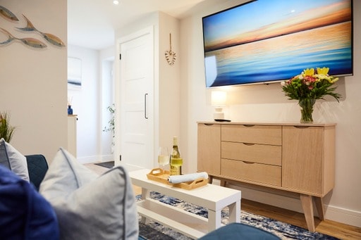A modern living area features a light wooden console table with a bouquet of flowers and a bottle of wine. A large television displays a tranquil seascape image. Soft blue cushions adorn the dark sofa, and light streams in from a nearby doorway, brightening the space.