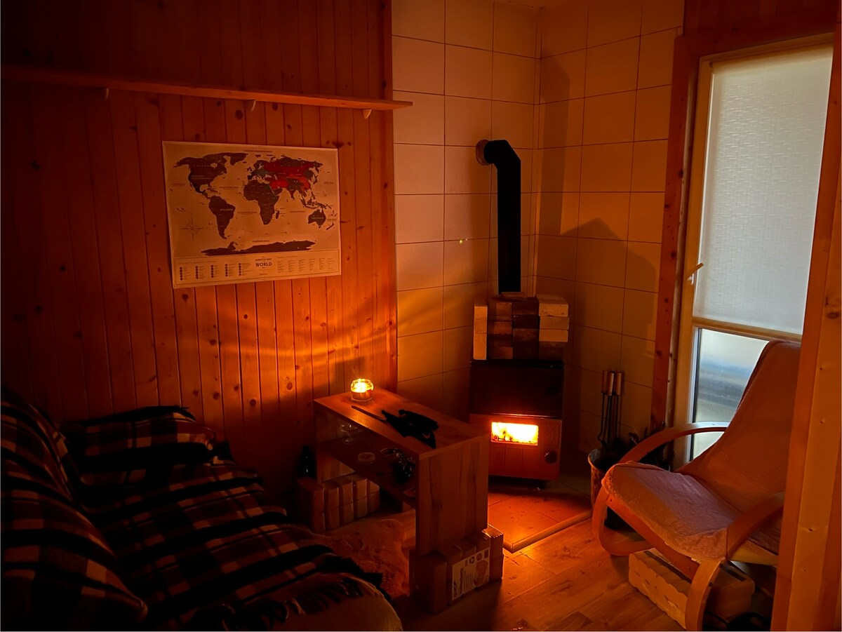 A warm corner features a wooden wall and a cozy couch covered with a checkered blanket. A softly glowing fireplace is positioned nearby, complemented by a simple table made of wooden crates. A world map hangs on the wall, adding a touch of adventure.