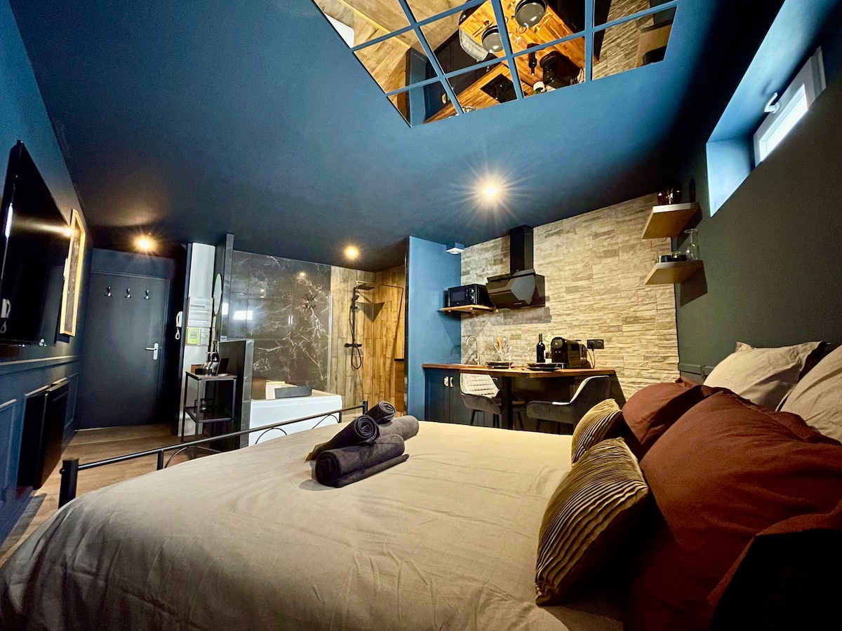 A modern room features a comfortable bed adorned with plush pillows, framed by dark blue walls. The kitchenette with sleek appliances is partially visible, along with a stylish bathroom area featuring a walk-in shower. Soft lighting enhances the inviting atmosphere.