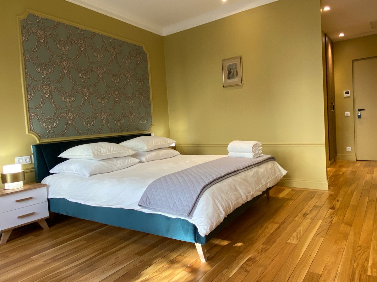 A spacious bedroom features a large, well-made bed with multiple pillows and a decorative headboard. Natural light highlights the wooden flooring and soft green walls, while a small bedside table with a lamp adds functionality to the space.