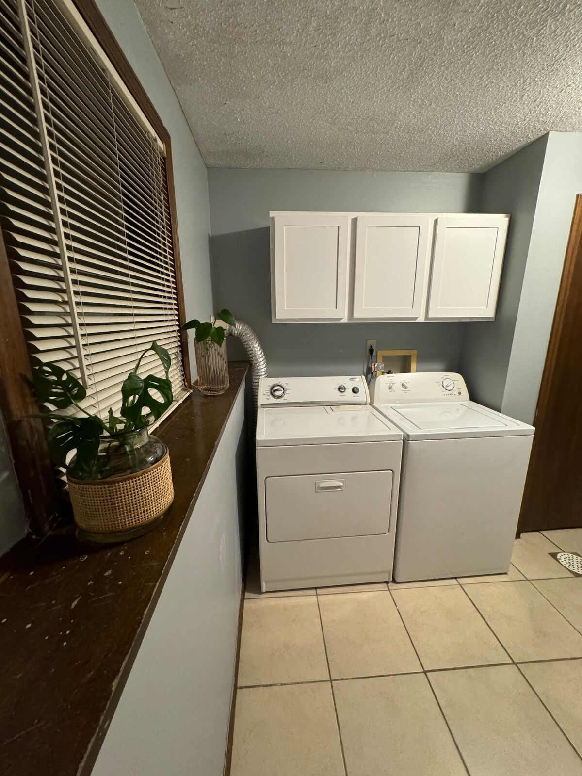 A laundry area is highlighted by a pair of white washers and dryers, situated beside simple cabinetry. A potted plant adds a touch of green, while a window with neatly aligned blinds allows soft light into the space. Neutral-colored tiles complete the functional setting.