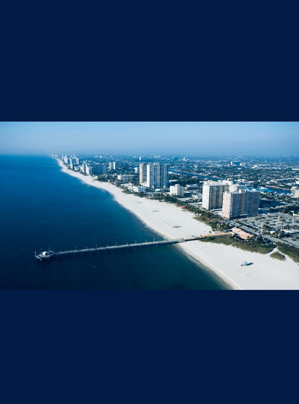 An aerial view captures the coastline featuring a long, sandy beach meeting calm waters. Skyscrapers line the shore, with a wooden pier extending into the water. The skyline includes distant buildings, creating a blend of natural and urban elements.