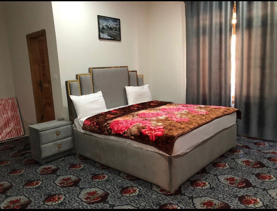 A spacious sleeping area is presented, featuring a large bed with a floral-patterned blanket and two upholstered pillows. Soft grey decor complements the patterned carpet below. Natural light filters through floor-length curtains, enhancing the inviting ambiance.