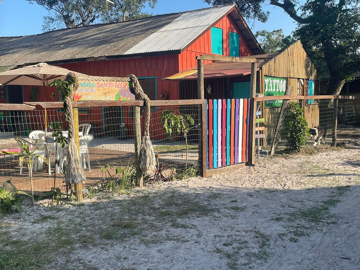 A vibrant exterior of a rustic building is surrounded by sandy terrain and greenery. The entrance features a colorful striped gate, while a patio area with white chairs and umbrellas is visible. The warm tones of the red and wooden structures enhance the welcoming atmosphere.