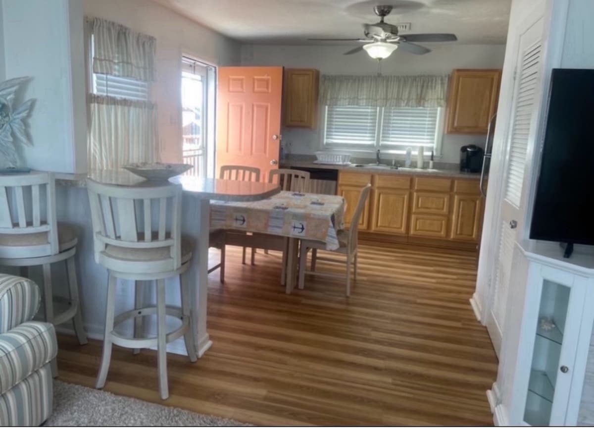 An open kitchen and dining area features light wood cabinetry and a dining table surrounded by four chairs. A breakfast bar with two stools lines the edge of the kitchen. Natural light filters through the window, enhancing the warm wood flooring.