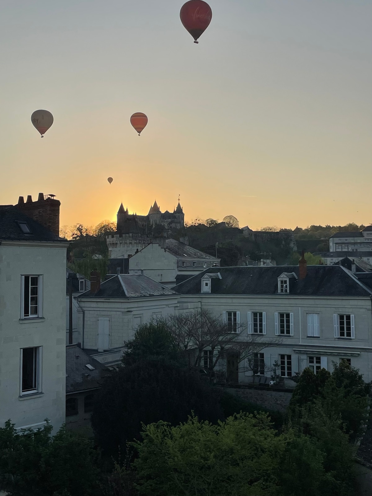 Colorful hot air balloons rise against a soft orange sky, with the silhouette of Saumur Castle in the background. The scene captures a tranquil morning, framed by rooftops and greenery below, suggesting a beautiful start to the day.