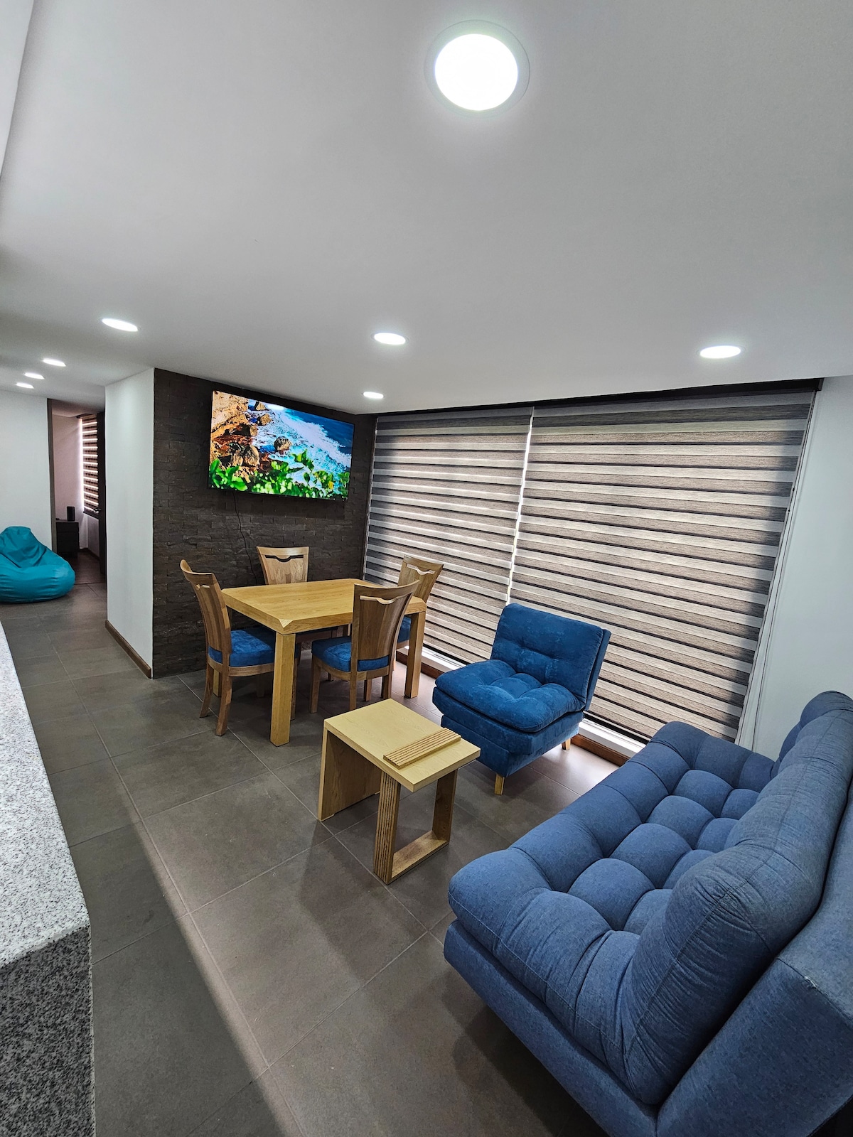A modern living area is presented with a blue sofa, a small wooden table, and four dining chairs surrounding a square table. A large wall-mounted TV displays vibrant imagery. Natural light filters through stylish window blinds, illuminating the space.