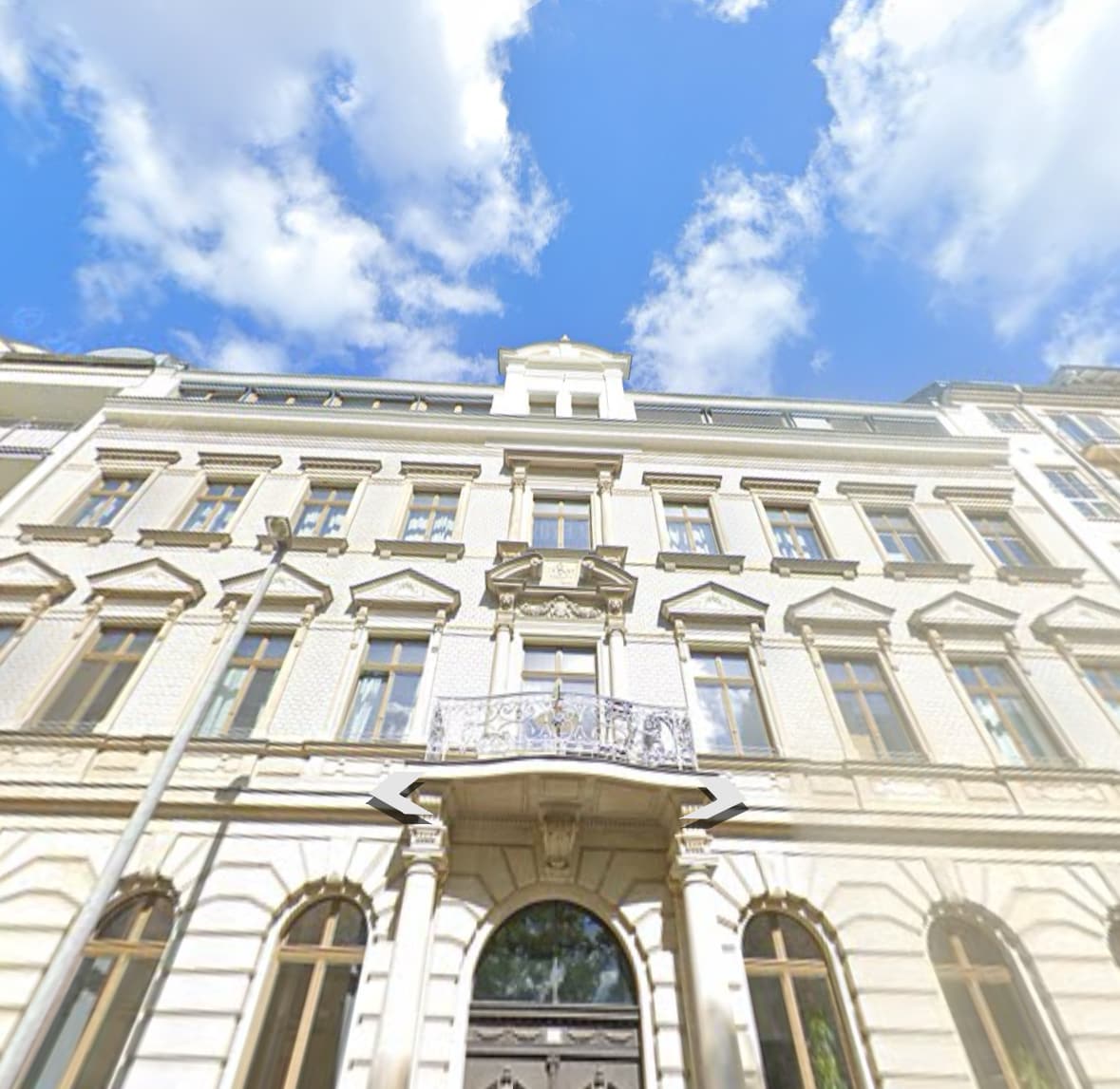 The historic building features an elegant façade with intricate architectural details. Large windows allow natural light to fill the interior, and a decorative balcony enhances the structure's charm. A clear blue sky provides a striking backdrop, complementing the overall aesthetic.