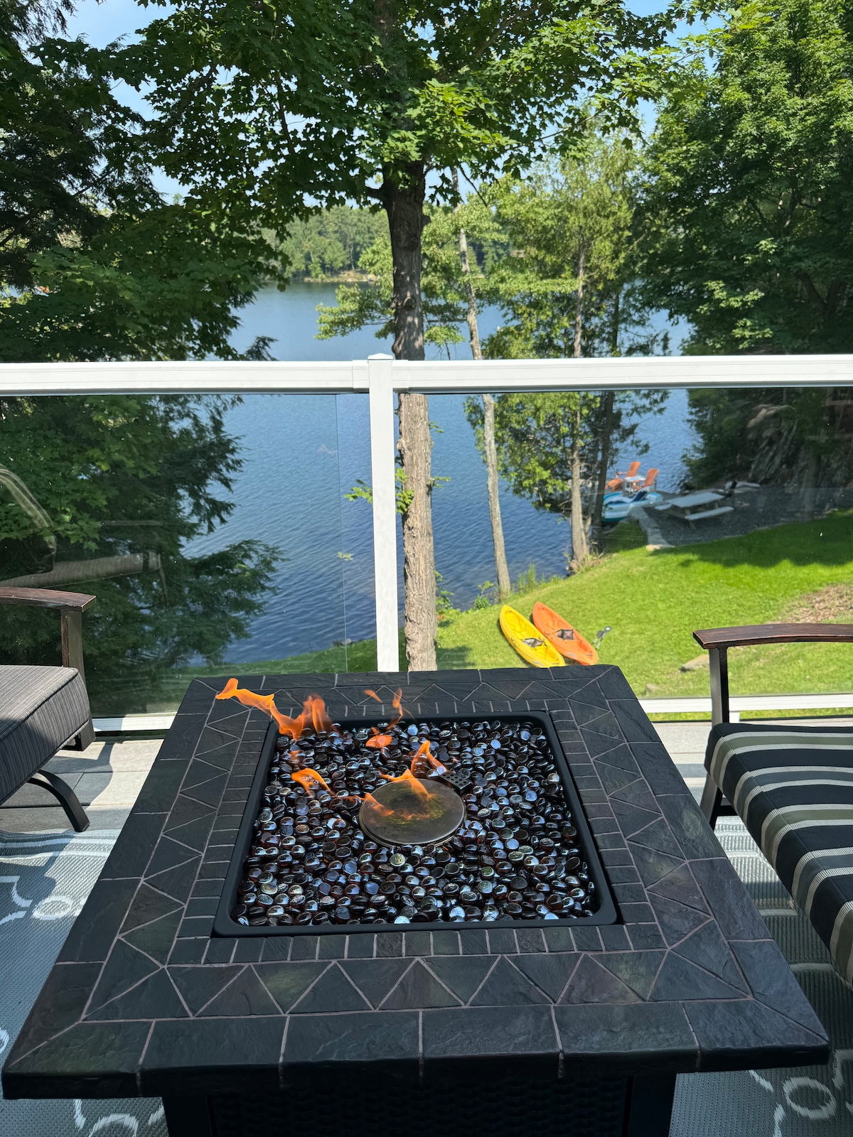 A fire pit with glass beads is centered on a balcony, providing warmth and ambiance. The calm lake is visible in the background, surrounded by trees. Two kayaks are positioned by the water's edge, ready for use, while comfortable seating invites relaxation.