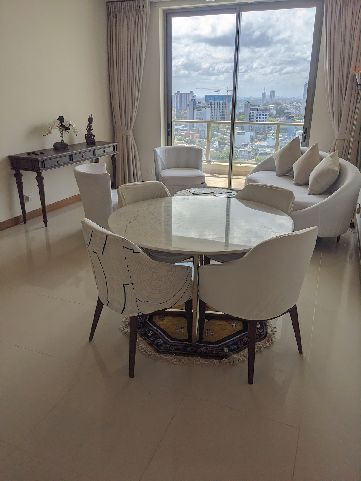 A dining area is presented, featuring a round marble table surrounded by four upholstered chairs. A comfortable armchair is positioned nearby. Large windows provide a view of the city skyline, with natural light brightening the space. Decorative pieces are visible on the console table.