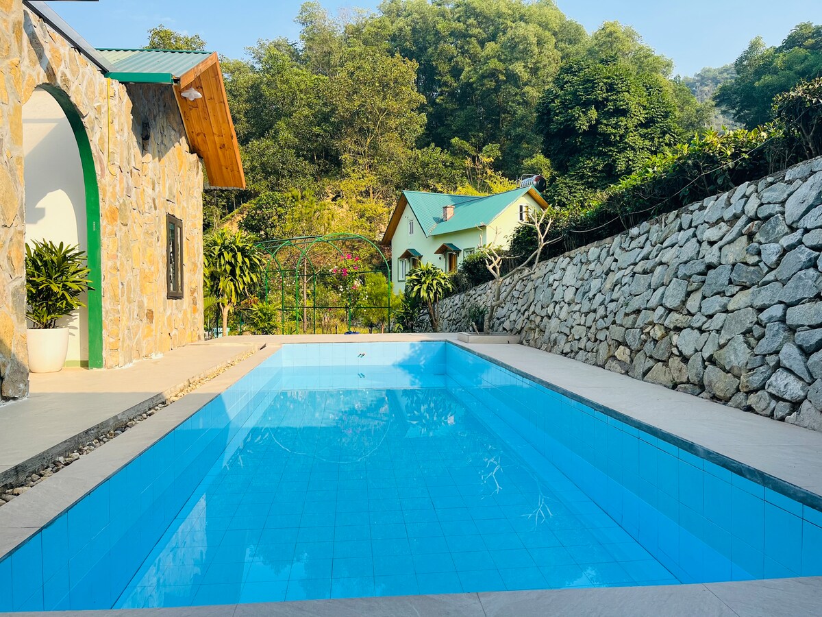 A serene swimming pool is surrounded by a stone wall and lush greenery, reflecting clear blue water. Two wooden-roofed cottages are visible in the background, nestled among the trees, contributing to a tranquil setting.