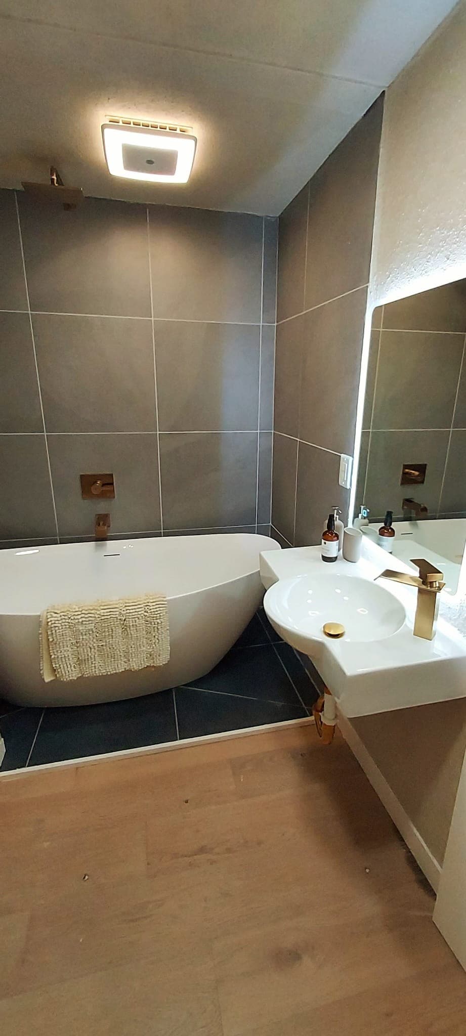 A modern bathroom featuring a freestanding bathtub with a textured towel draped over the side. The sleek, minimalist design is complemented by gray tiled walls and a light wood floor. A contemporary sink with gold fixtures is positioned next to a wall-mounted mirror.