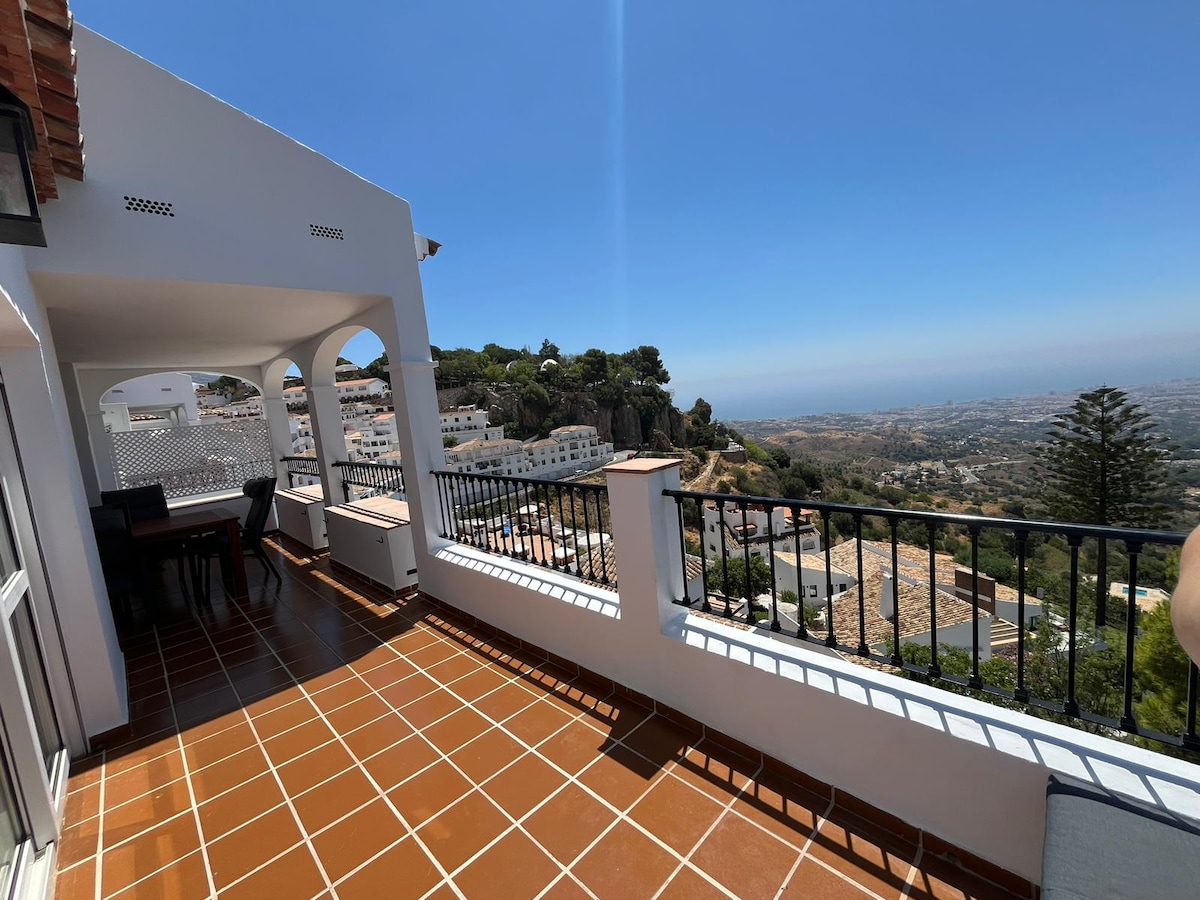 A spacious terrace features terracotta tile flooring and a metal railing, offering panoramic views of the surrounding landscape. Arched openings frame the outdoor area, which includes a dining table set for four. Sunny blue skies complement the serene atmosphere of this elevated space.