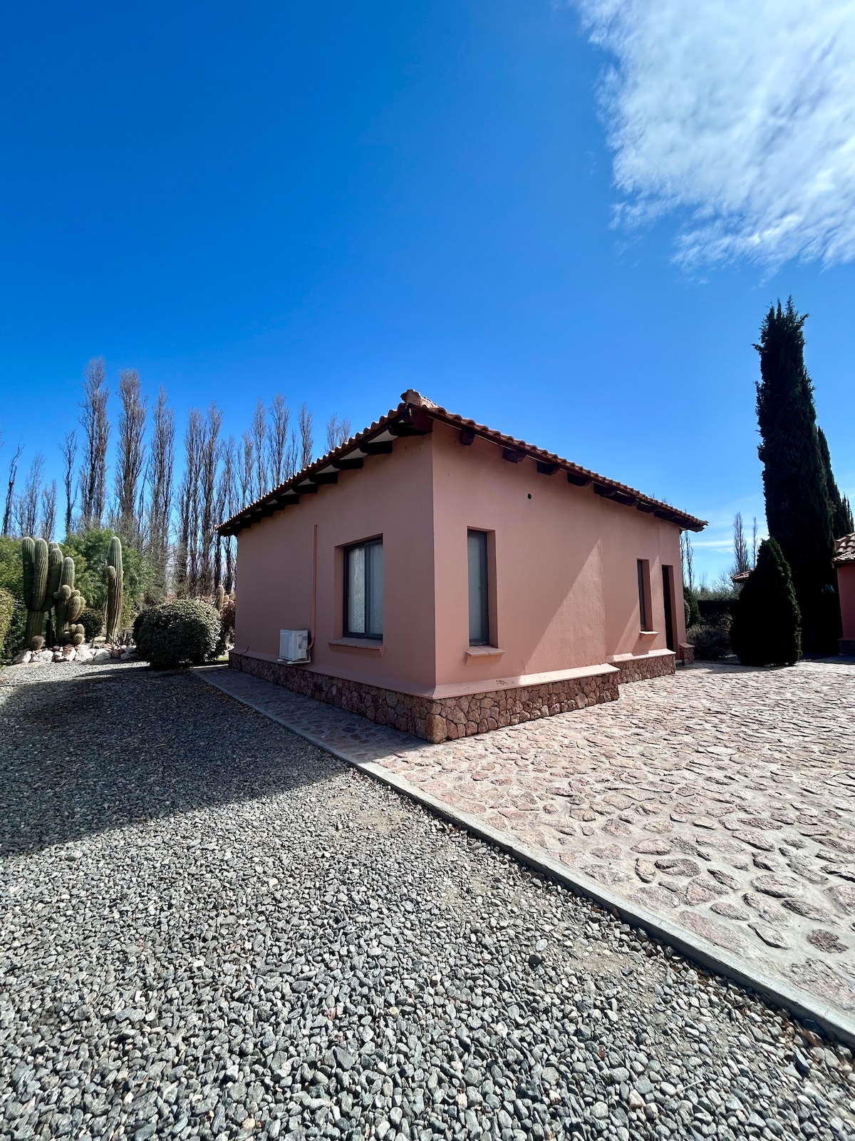 A single-story building with a soft pink exterior is shown against a bright blue sky. The structure features a textured stone pathway and is surrounded by landscaped greenery, including tall trees and cacti, emphasizing a serene and natural environment.