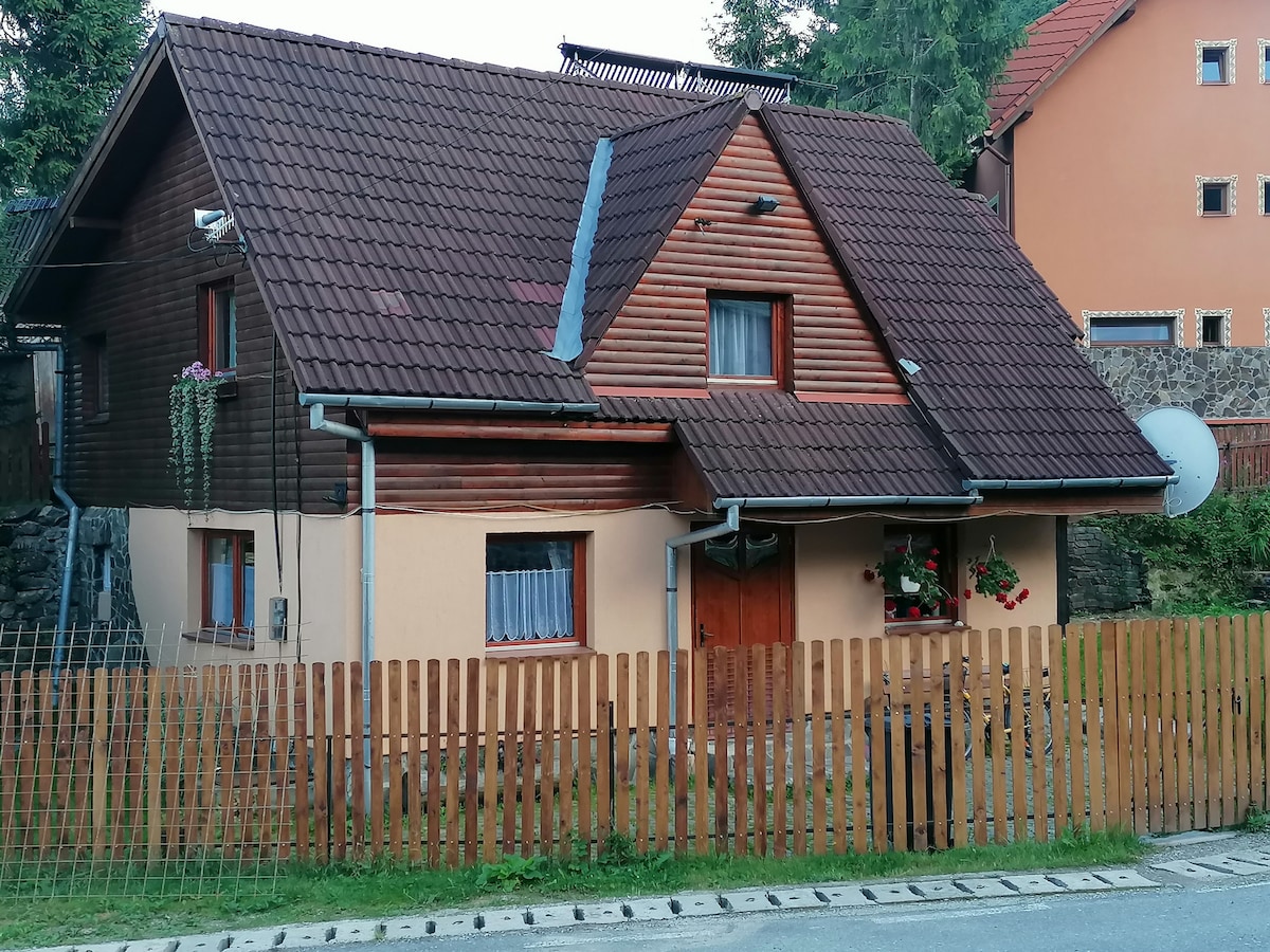 A two-story house features a mix of wooden and painted exterior walls. The sloped roof is covered with dark tiles, and flower boxes adorn the windows. A wooden fence encloses the front yard, and a pathway leads to the door, creating a welcoming entrance.