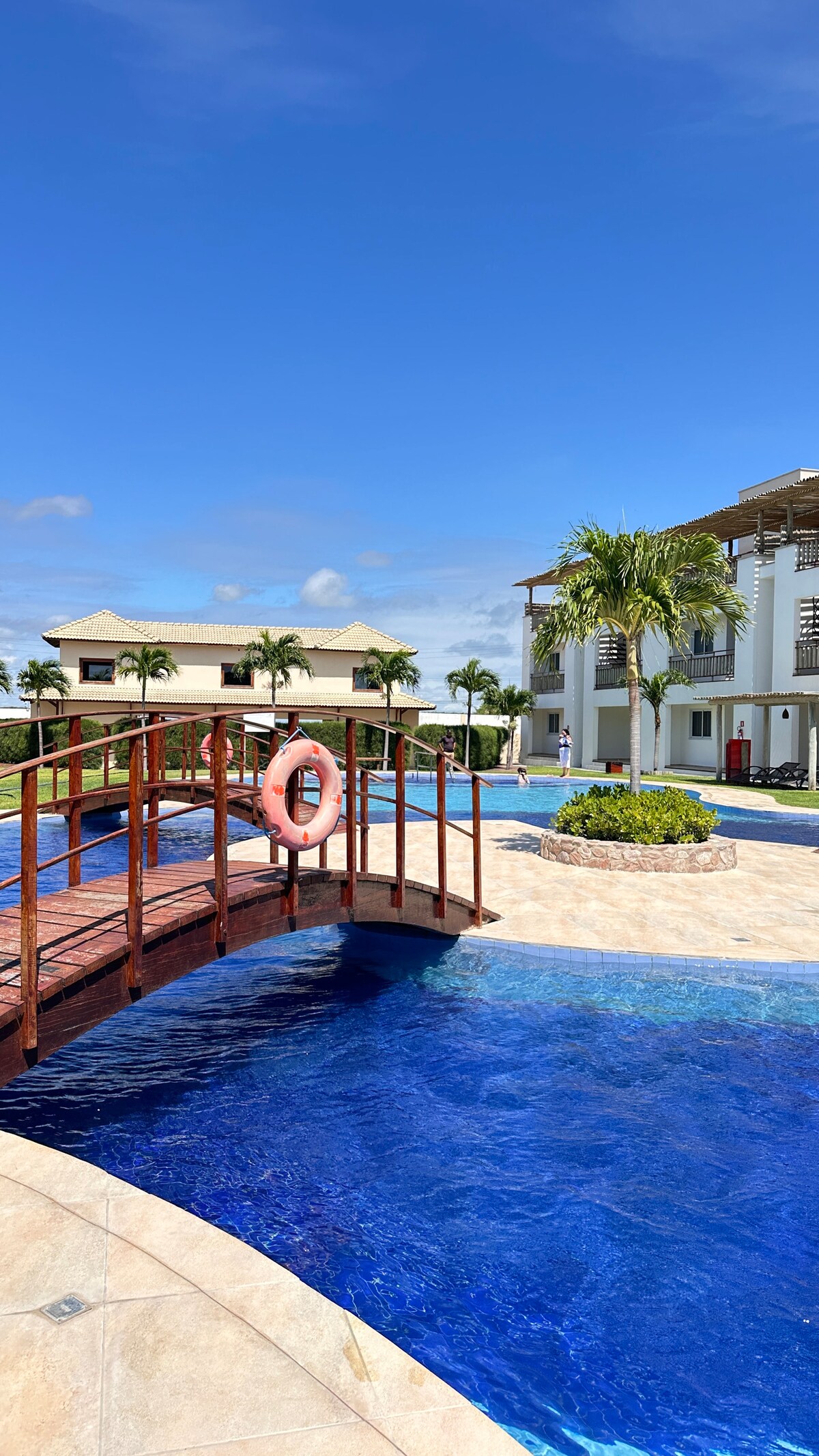 A wooden bridge with a lifebuoy spans a sparkling blue pool surrounded by lush greenery. Palm trees and attractive buildings are visible in the background, contributing to a serene outdoor atmosphere. Clear skies and ample sunlight enhance the tranquil setting.