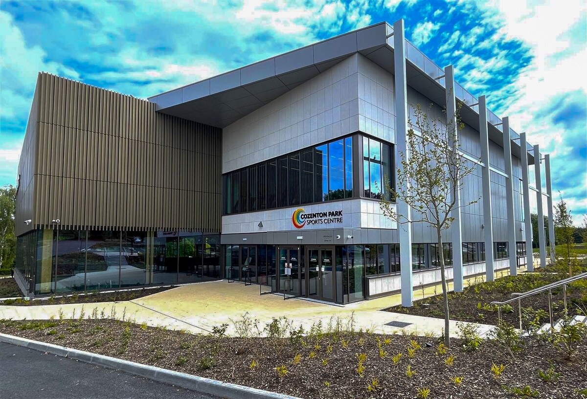 The exterior of a modern sports center is showcased, featuring a combination of light and dark grey paneling. Large glass windows line the facade, allowing natural light into the building. Surrounding landscaping includes a paved walkway and small trees.