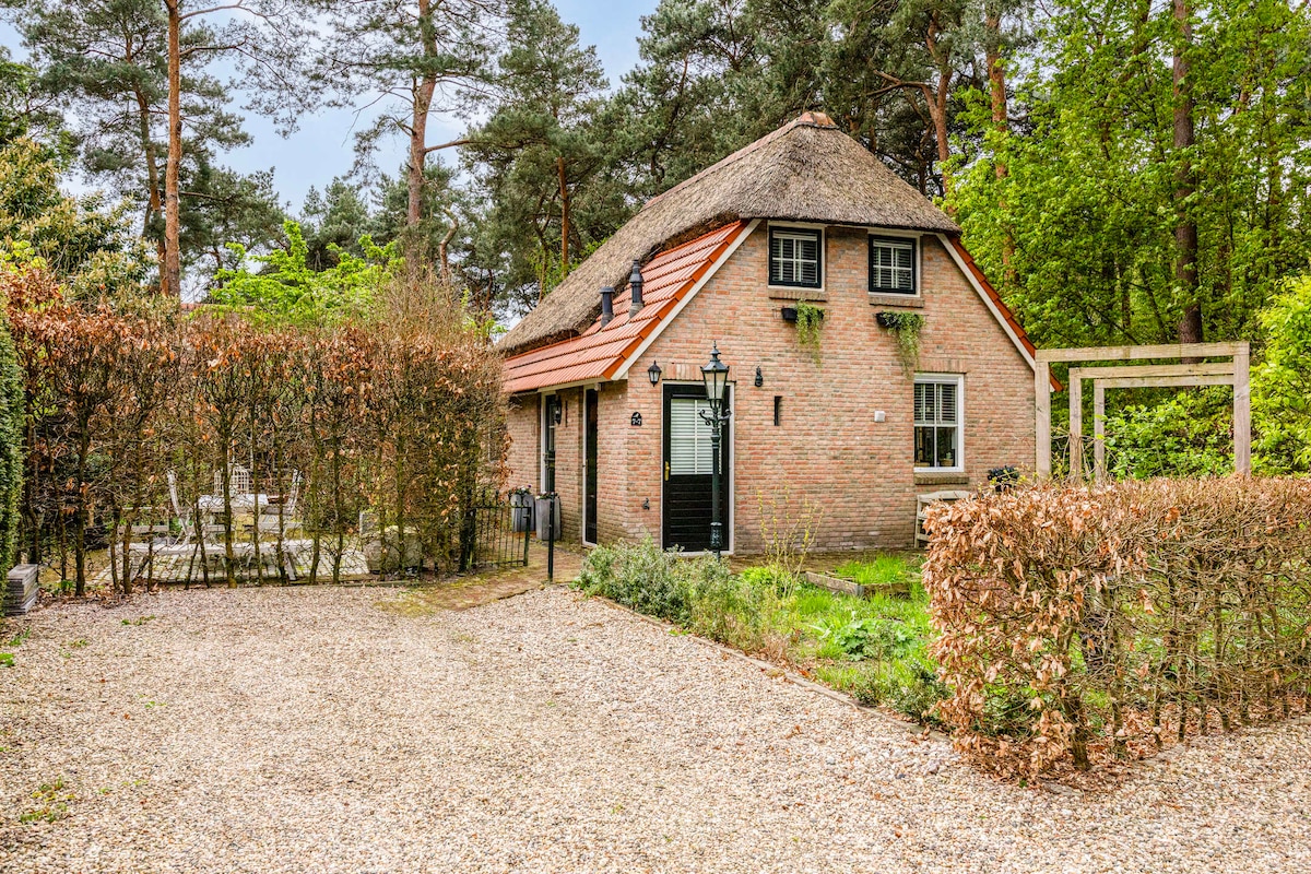 A charming vacation home with a thatched roof is nestled among trees. The exterior is surrounded by a well-maintained garden and a hedge. A gravel driveway leads to the entrance, providing a sense of privacy in the natural setting.